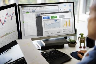 Business professional at desk with two monitors that display Microsoft excel spreadsheets, graphs, and tables